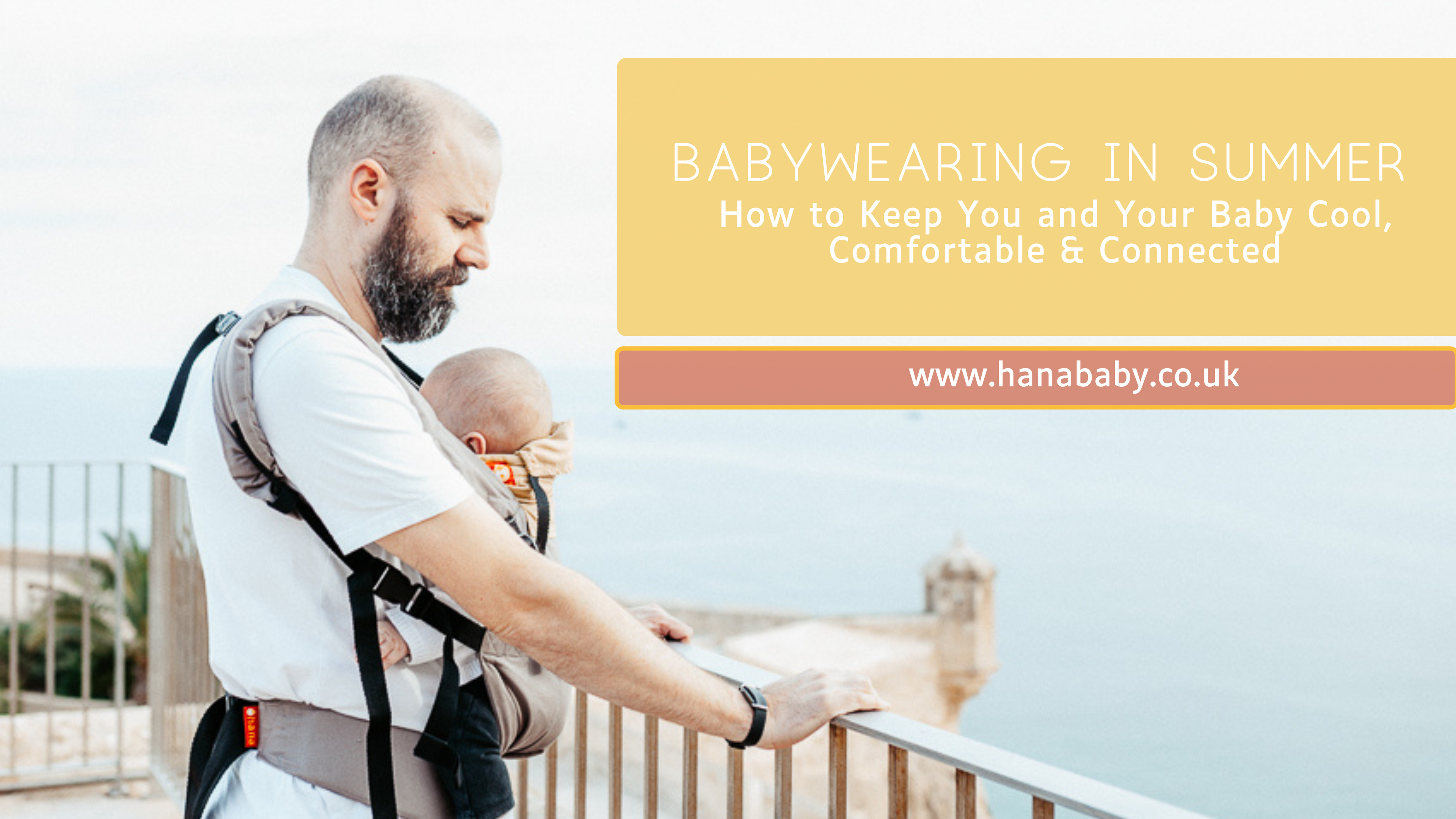 A Daddy on holiday stands against railings over looking the sea from Alicante Castle. He is wearing his three month old baby in the Hana Baby Expand, which is grey and yellow