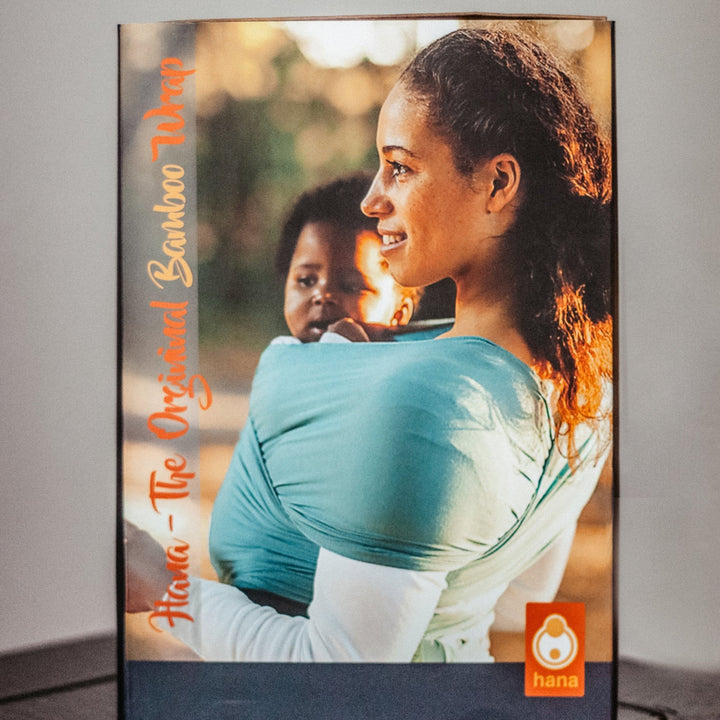 Woman holding a child in a baby carrier with 'hana' branding on a gray background demonstrating the perfect baby gift