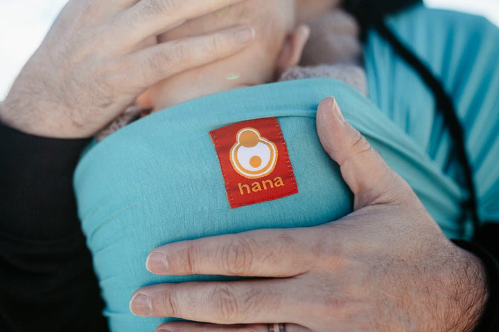 A Fathers hands are shown supporting his baby whilst showcasing the hana baby logo against a light blue baby carrier. Hana Baby makes organic baby carriers for newborns.