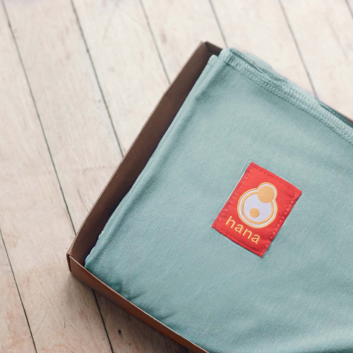 Teal fabric sling with an orange 'hana' logo on a wooden surface. A great new baby gift.