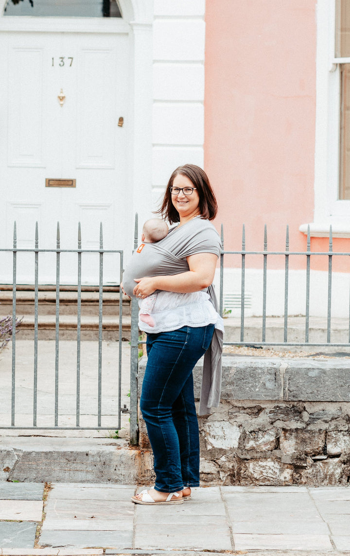 A mother wears her daughter is a light grey baby sling on a victorian street. She smiles as she walks.