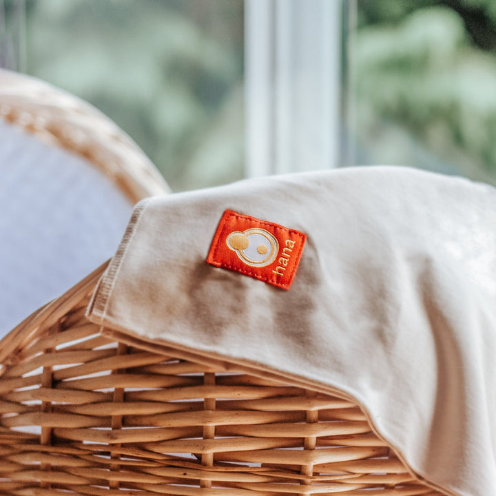 Wicker Moses basket with a Hana Baby product draped across. The orange tag featuring the brand 'Hana' in a blurred outdoor setting. The perfect present for a new baby.