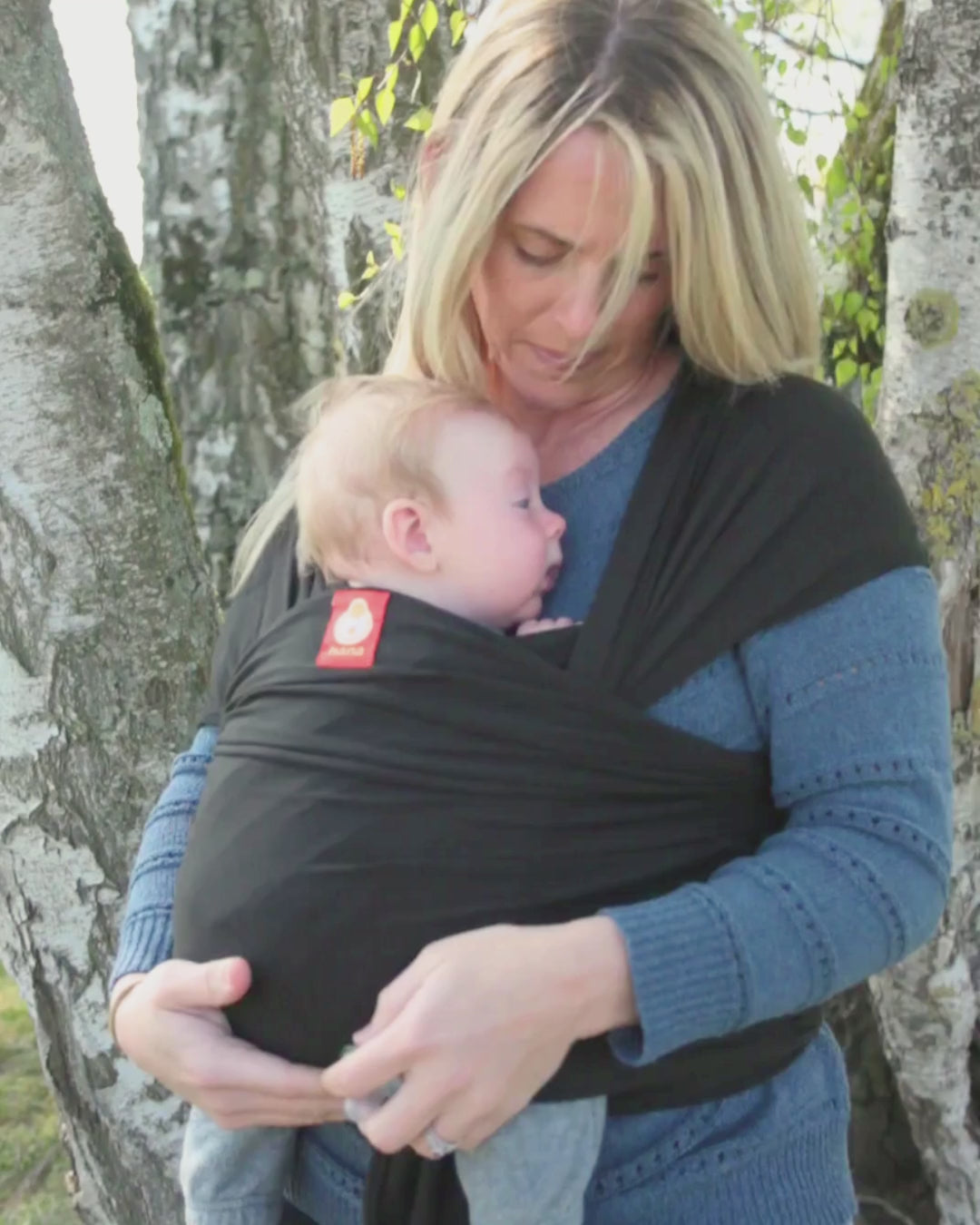 A mother carries her baby in a Hana Baby wrap carrier, a tree gentle blows in the wind as she cuddles her son in a black baby wrap.
