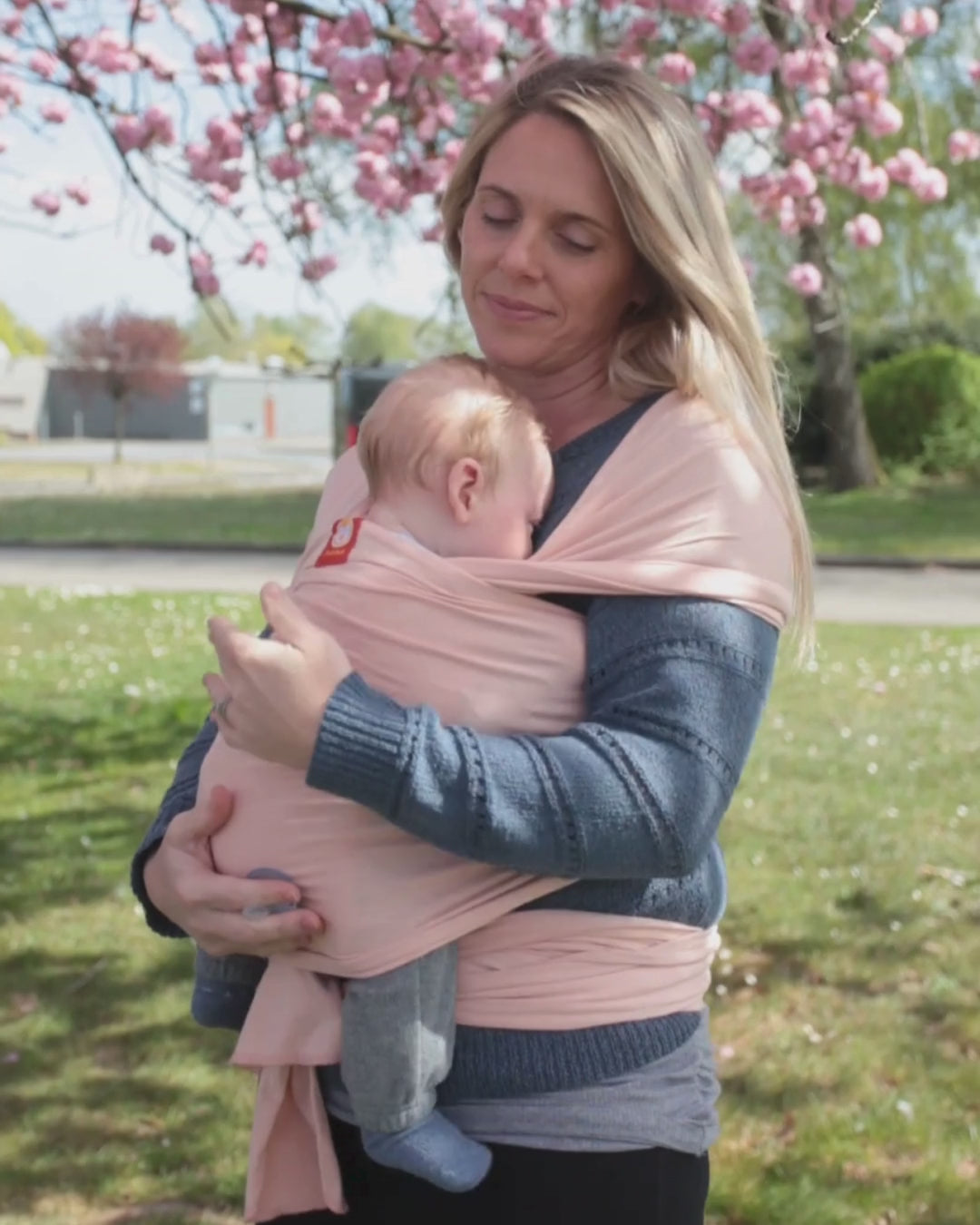 A mother cuddles her son in a light pink sling for newborns while she gently rocks him under blue skies and a blooming cherry blossom tree.