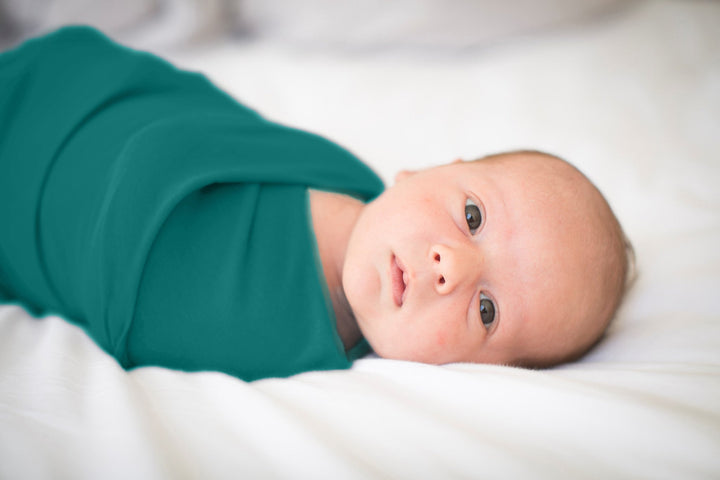Newborn baby wearing a teal baby blanket is lying on a white surface