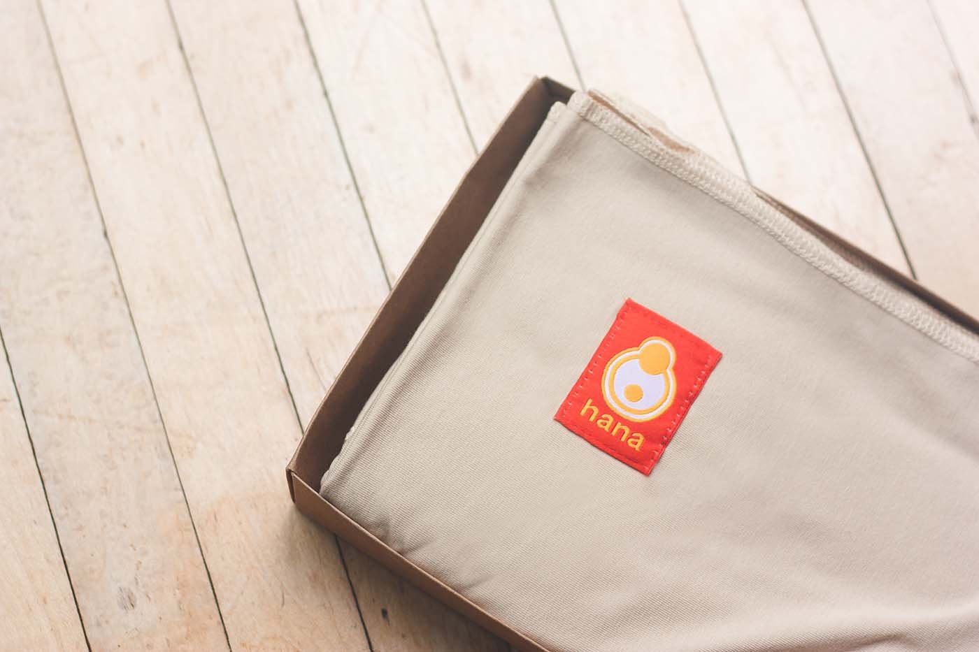 The image shows the Hana Baby Standard sling in the colour tan. Neatly folded in its box, placed upon an old wooden floor.