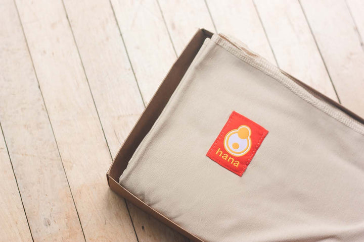 The image shows the Hana Baby Standard sling in the colour tan. Neatly folded in its box, placed upon an old wooden floor.