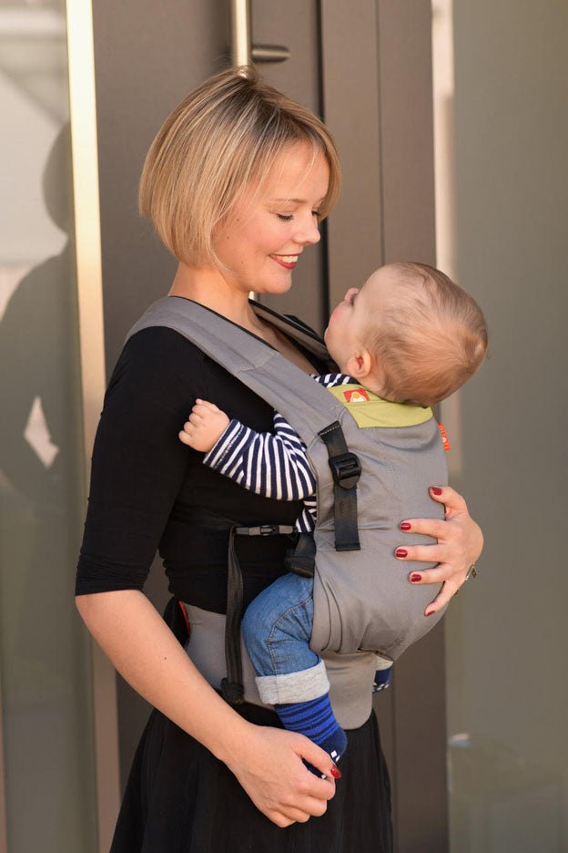 A mother stands in her doorway smiling down at her son who she is carrying in a grey and yellow hana baby expand baby carrier.