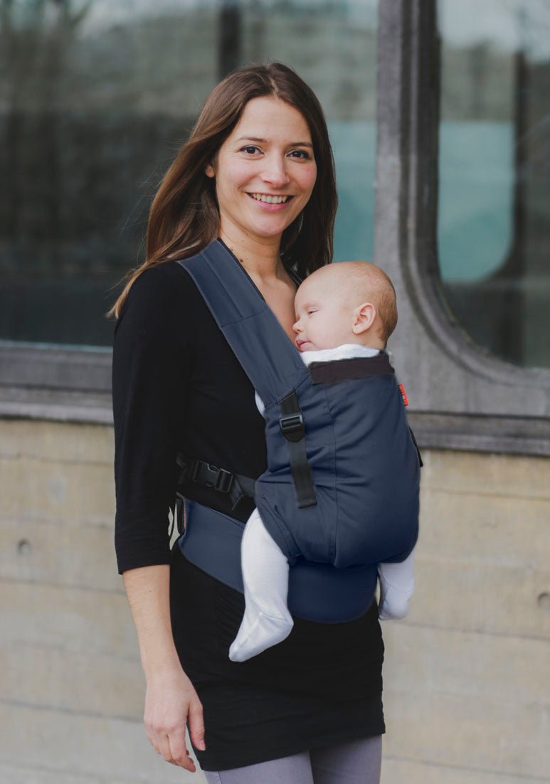 A mother smiles gently at the camera on an evening urban stroll. She carries her baby in a navy hana baby organic infant carrier.