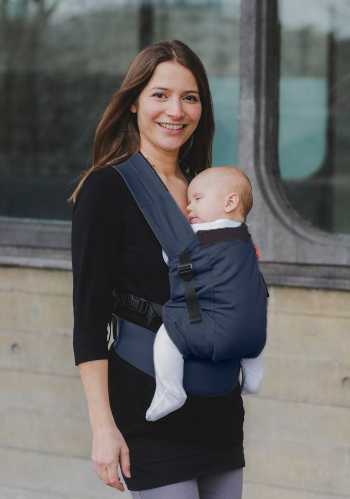 A mother smiles gently at the camera on an evening urban stroll. She carries her baby in a navy hana baby organic infant carrier.