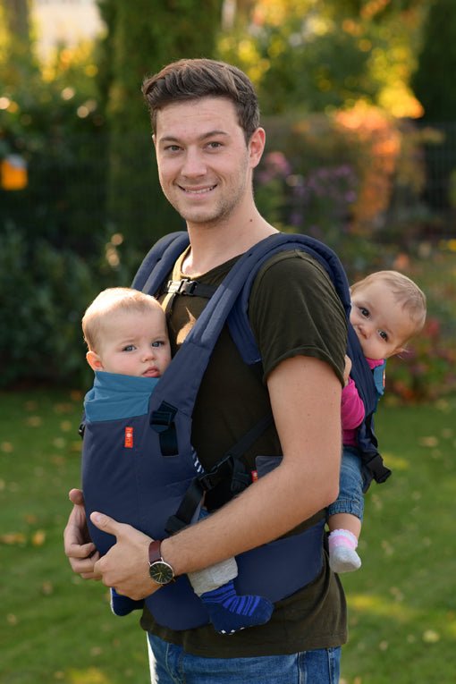 A father smiles in his garden as he carrier his son and daughter in the Hana Baby Expand twin baby carrier.