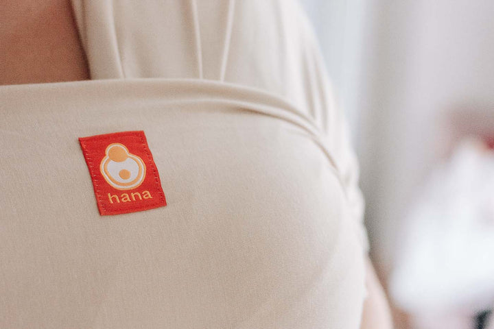 This image is a close up of the Hana Baby sling in tan. The focus of the image is the Hana Baby Logo.
