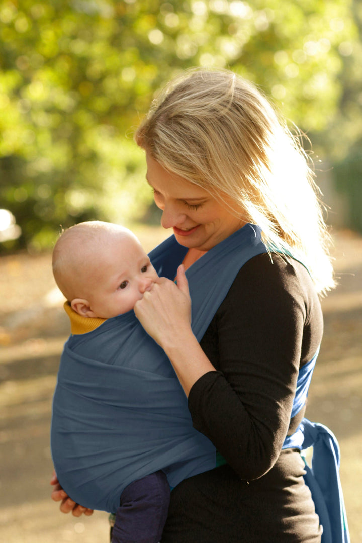 A mother gently smiles down at her baby who is watching the world go by snuggled in a navy baby sling wrap.