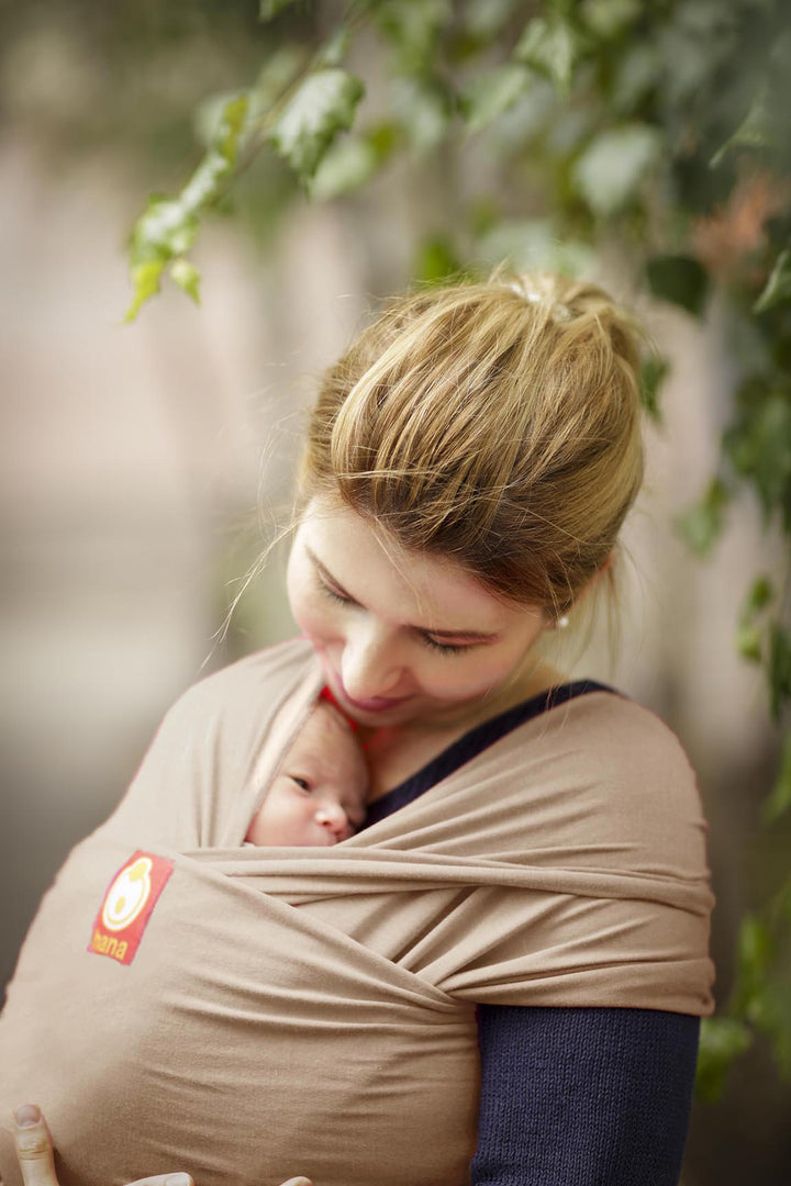 A mother glances down at her tiny baby peeking out from his baby sling wrap in a light tan colour.