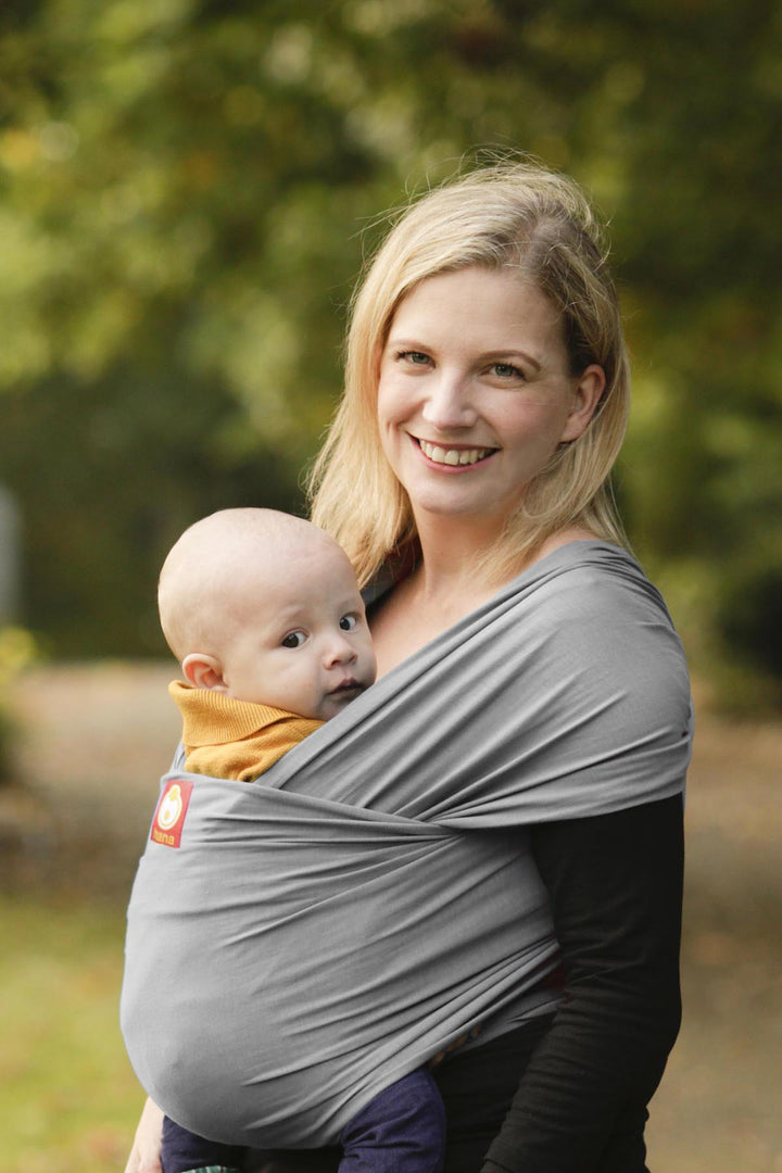 A mother smiles whilst she carries her baby in a light grey stretchy baby sling whilst strolling in a local park.