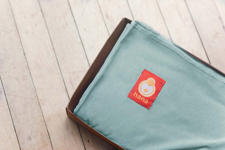 An images of a hana baby wrap in steel blue is neatly folded in a box sits upon an old school floor.