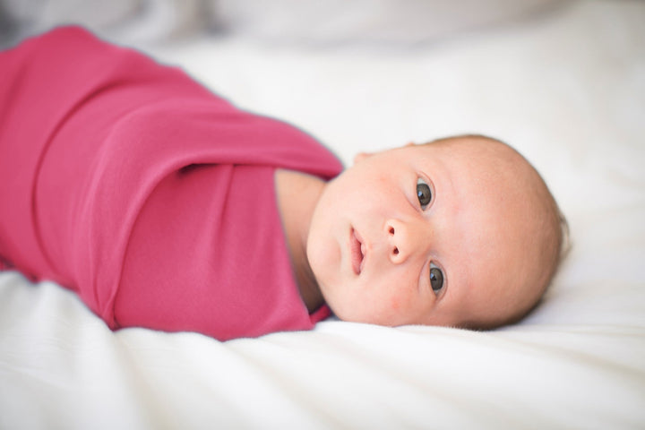 A Baby girls relaxes calmly on a white bed with her eyes fixed on the camera in a pink swaddle baby blanket