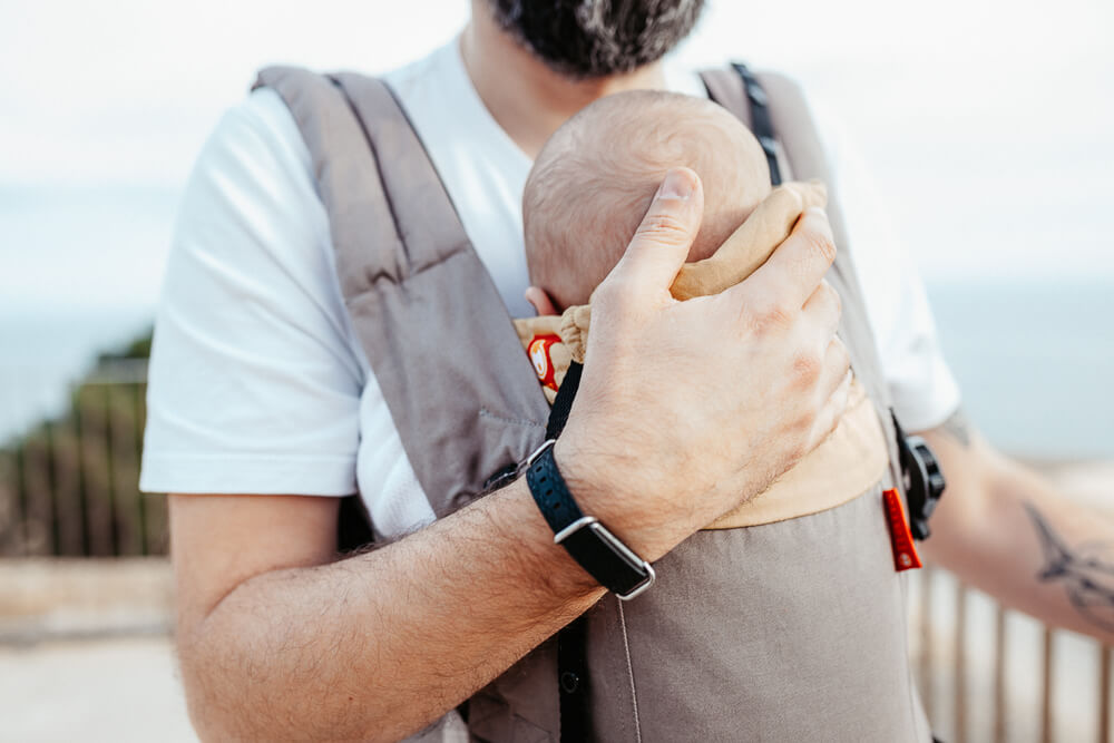 A father wearing a white top cradles his babies head whilst his baby sleeps in a grey baby carrier. In the background railings and a bush can be seen but these are blurred by a shallow depth of field. 