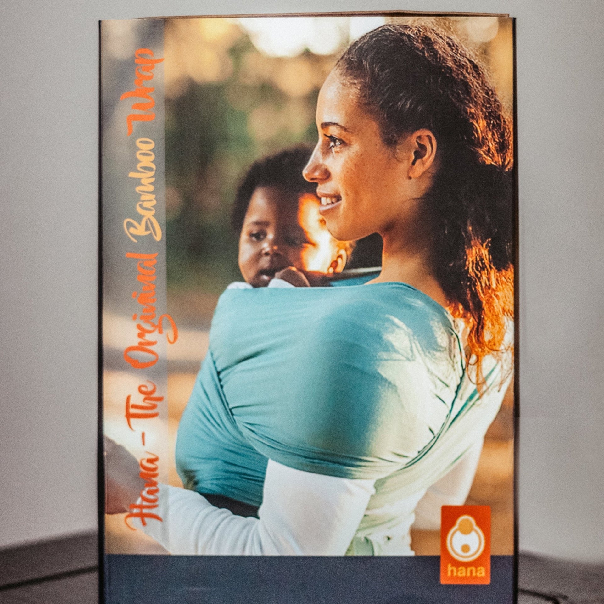 Woman holding a child in a baby carrier with 'hana' branding on a gray background demonstrating the perfect baby gift