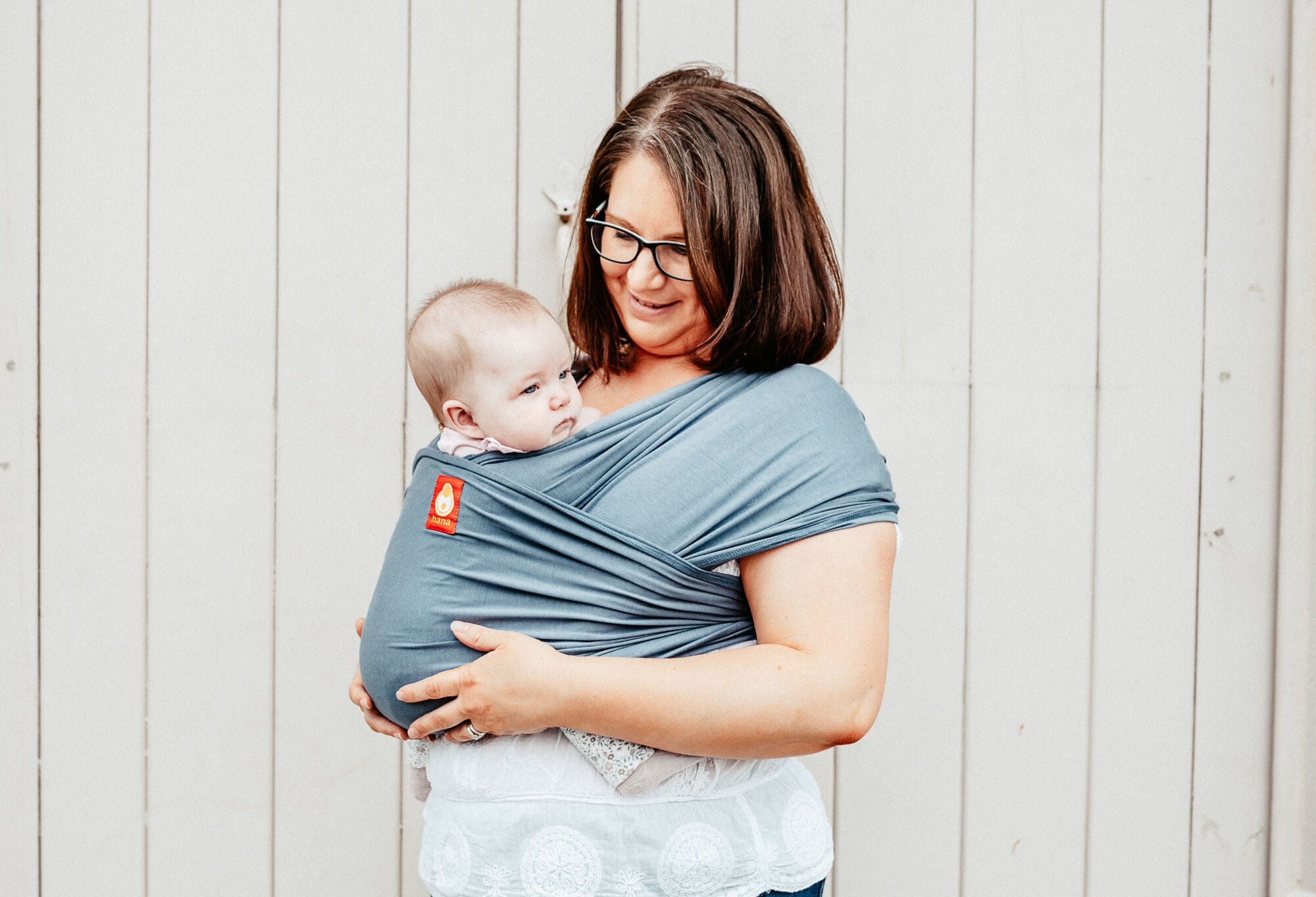 A mother looks Lovingly at her daughter who gases off into the distance safe, secure, and supported in a grey hana baby wrap. The pair stand against beige painted wooden doors. 